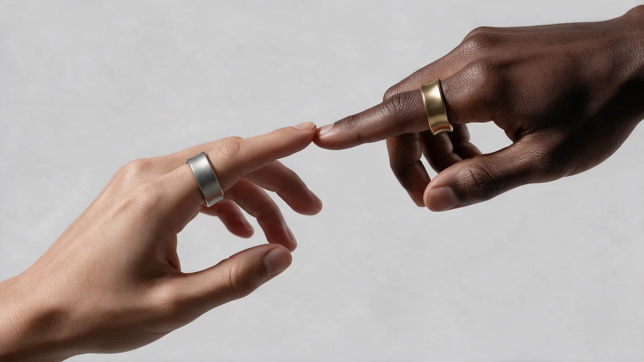 Two hands reach toward each other until the fingertips touch, one wearing the silver Ohm Ring and the other the gold